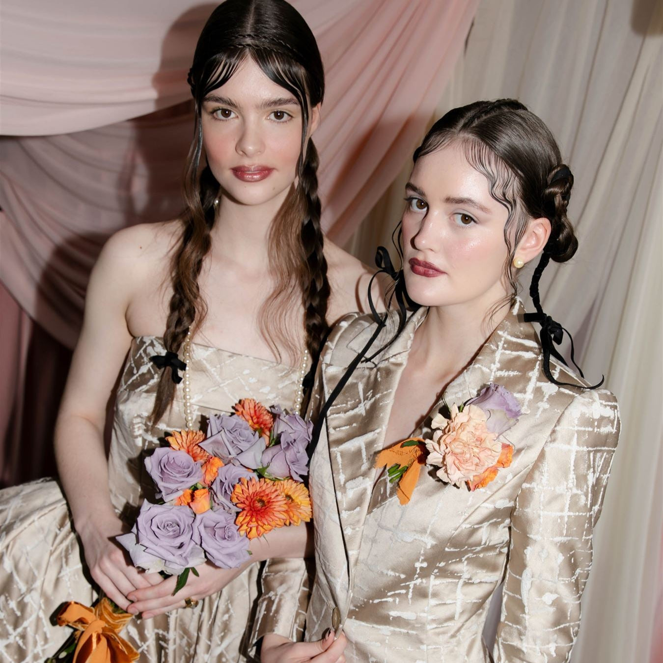 Two women in formal attire with floral bouquets standing against a draped curtain background.